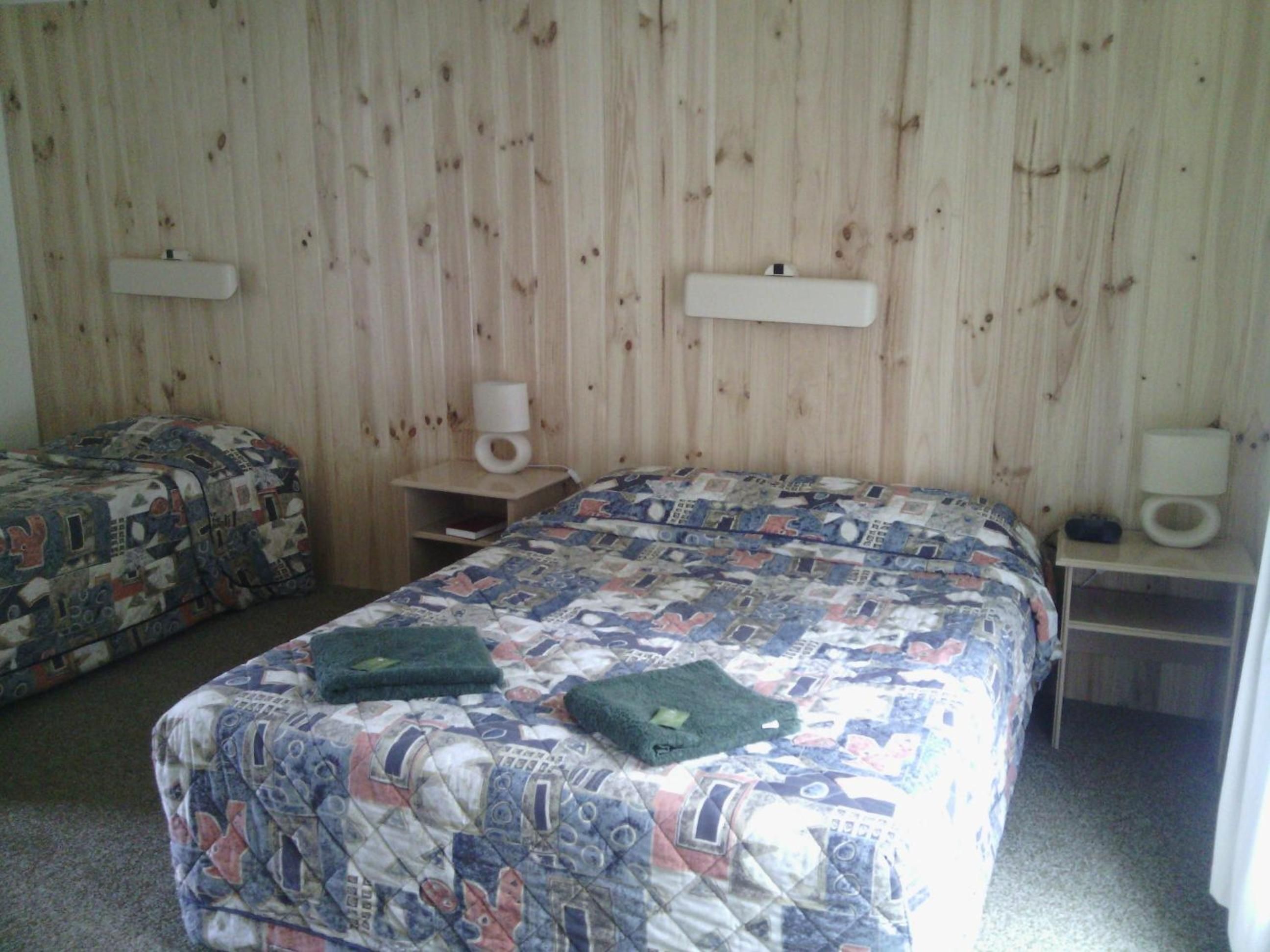 A room with two beds, each with patterned bedding, a bedside table with a lamp, and a wall-mounted shelf with a lamp.
