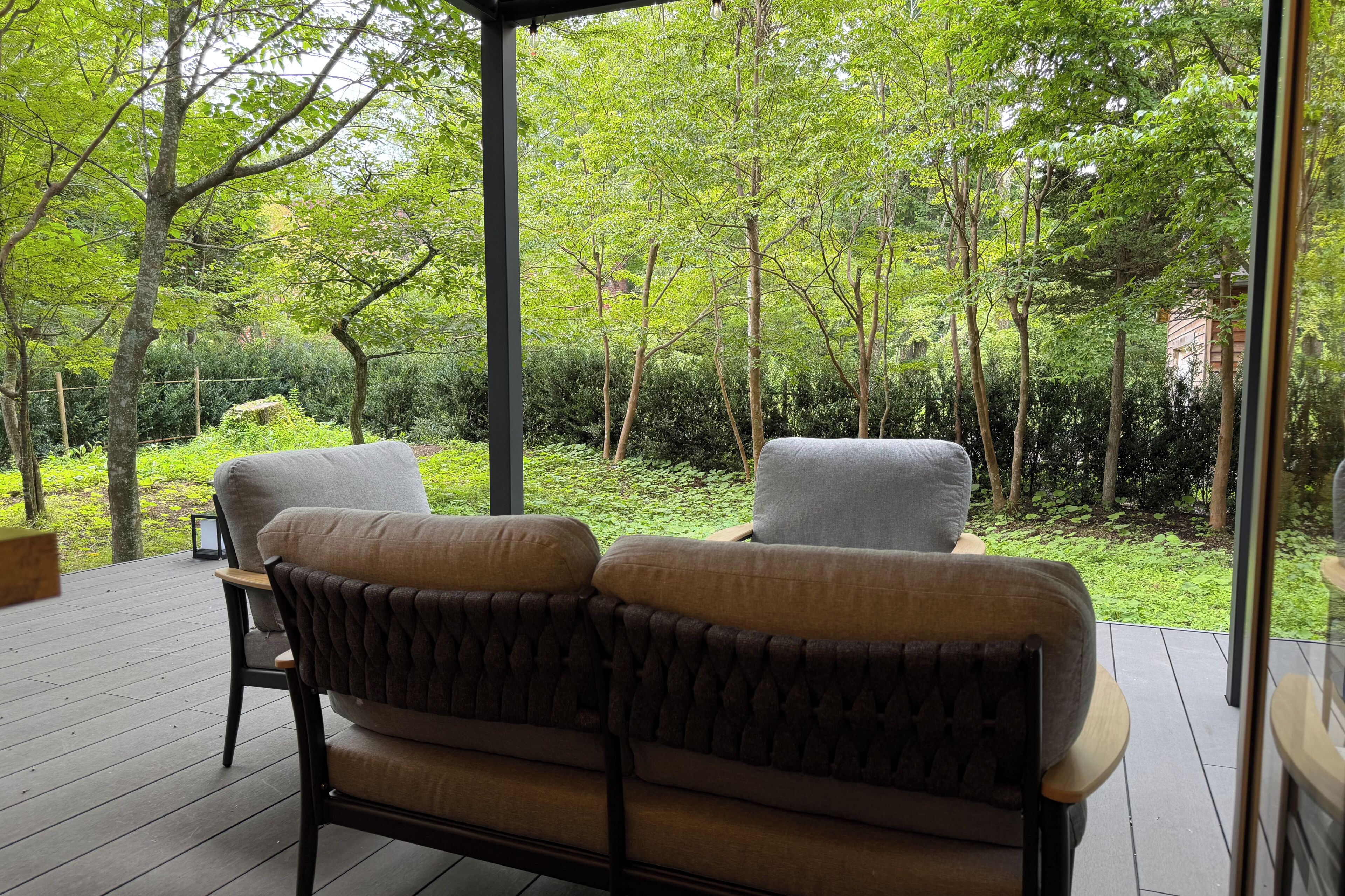 A covered outdoor seating area with two sofas and a table, surrounded by lush greenery and trees.
