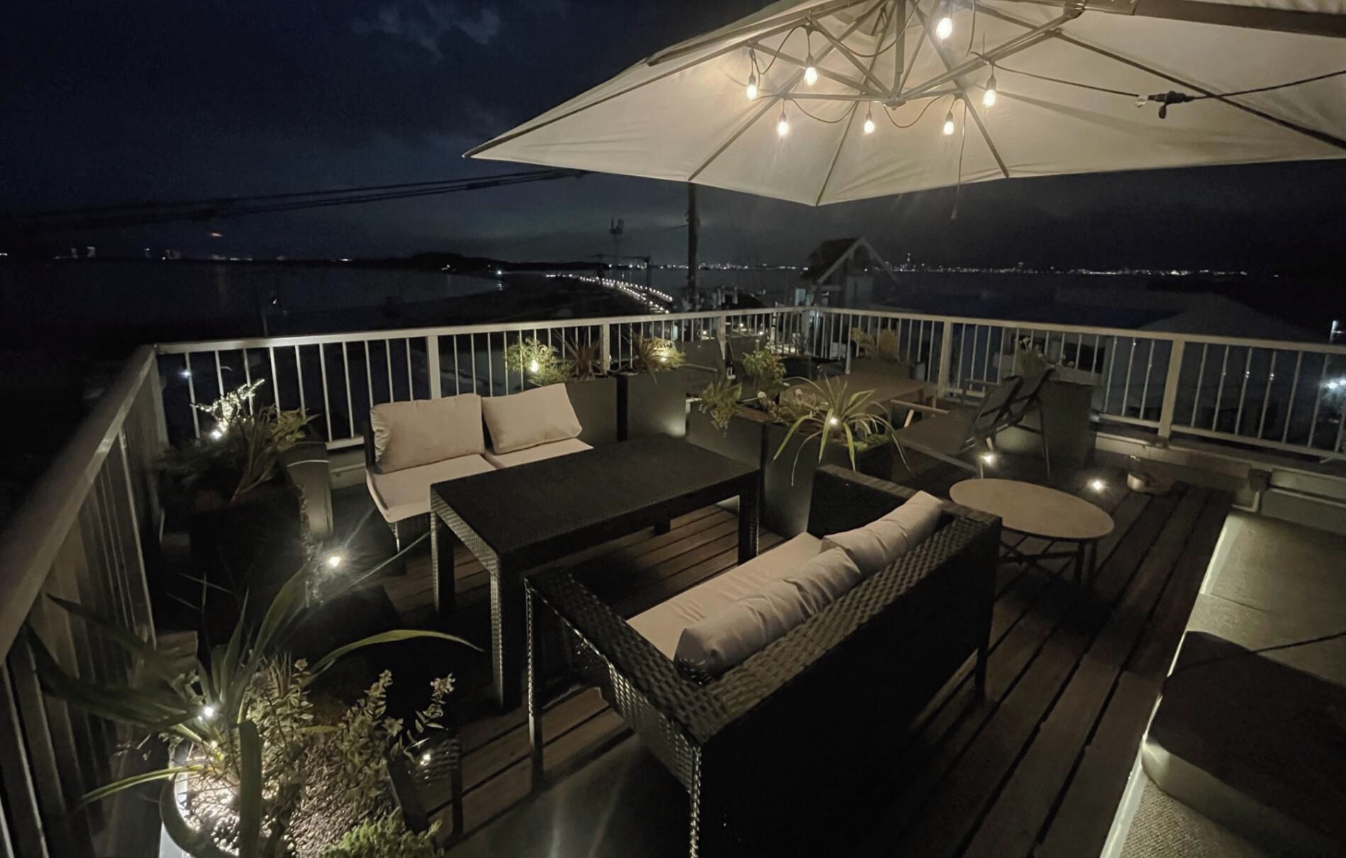 Eine Terrasse im Freien mit Esstisch, Sofa und Stühlen, umgeben von Blumentöpfen und einem großen Sonnenschirm.