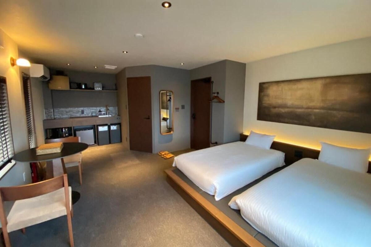 A hotel room with two beds, a desk, a chair, a small kitchenette, and a mirror.