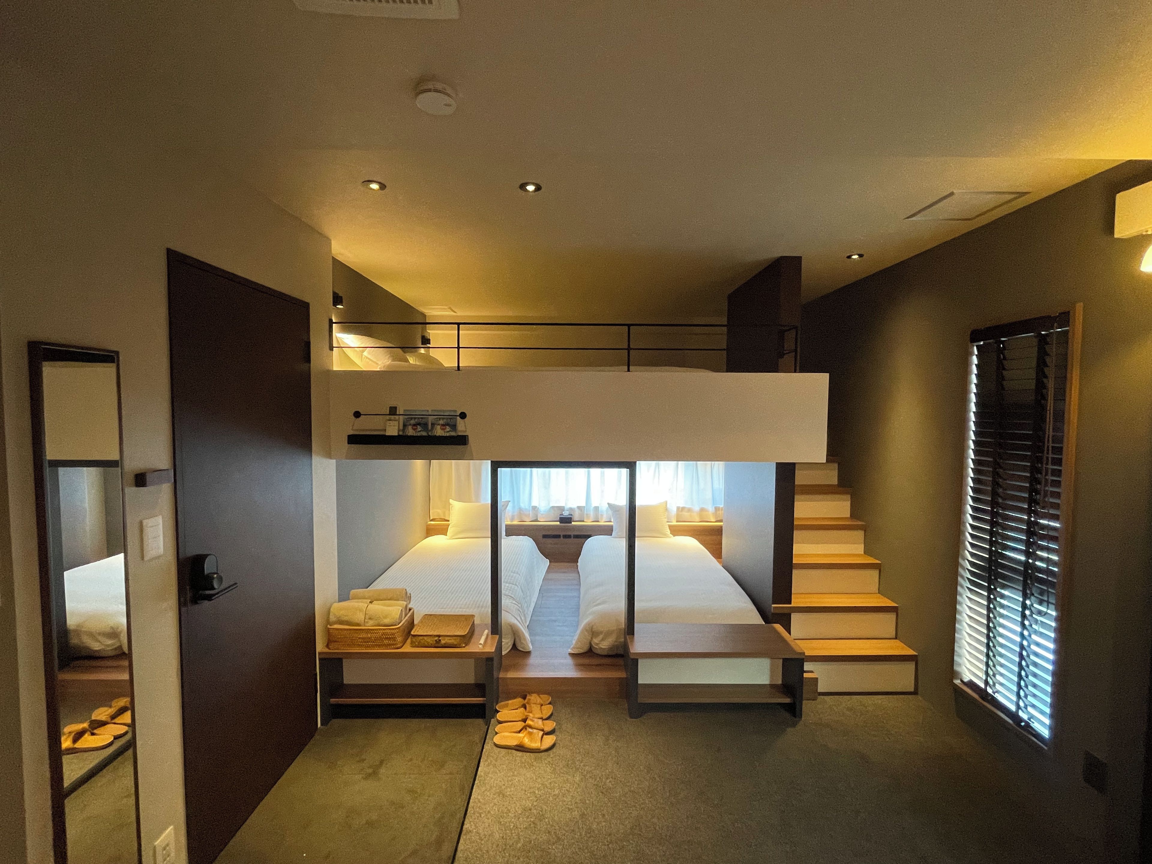 Ein modernes Hotelzimmer mit Etagenbett, einem kleinen Schreibtisch, einem Fenster mit Jalousien und einer Treppe.