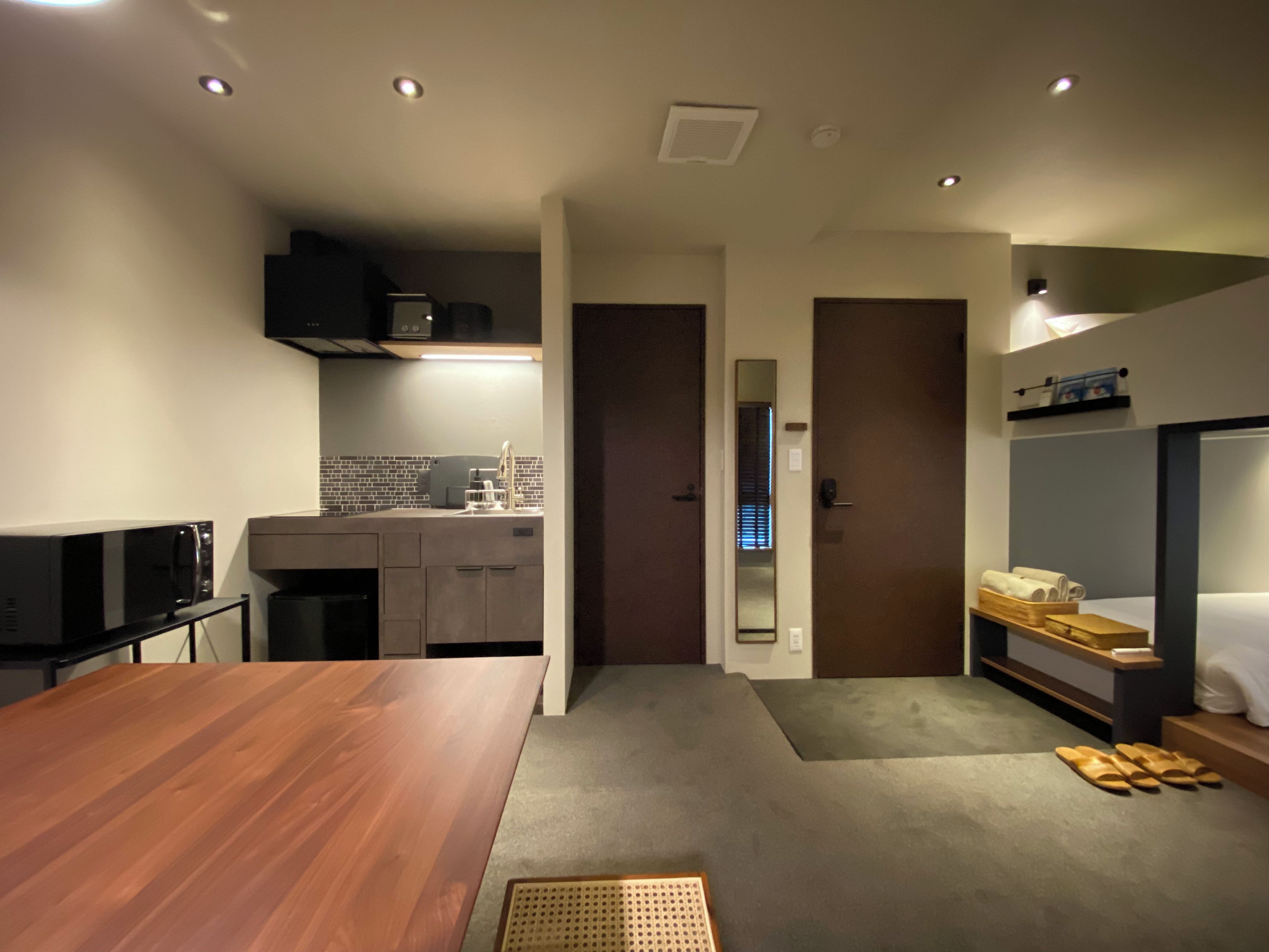 A compact hotel room with a kitchenette, a dining table, a bed with a wooden headboard, and a small seating area.