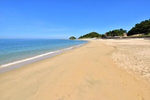 A beach with clear blue water, sandy shore, and a small hill in the distance.