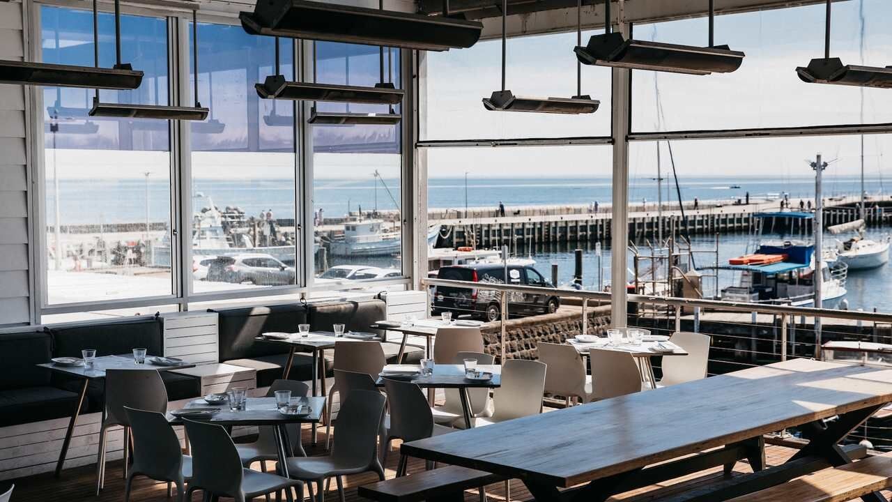 Un espace extérieur aménagé pour les repas, avec des tables et des chaises, offrant une vue sur une marina avec des bateaux et une jetée.