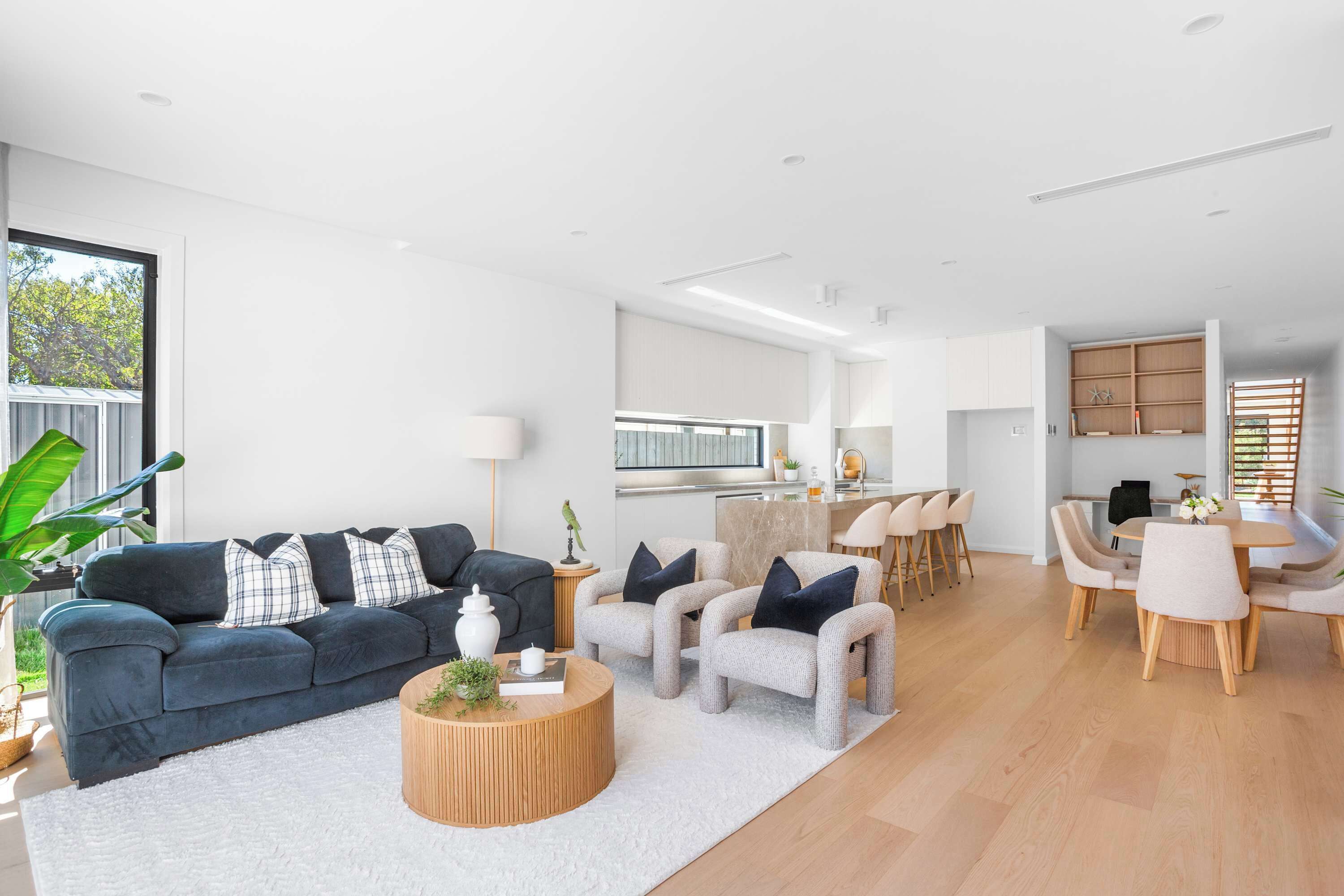 A modern living room with a dark blue sofa, light-colored armchairs, a wooden coffee table, and a kitchen area in the background.