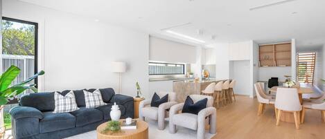A modern living room with a dark blue sofa, light-colored armchairs, a wooden coffee table, and a kitchen area in the background.