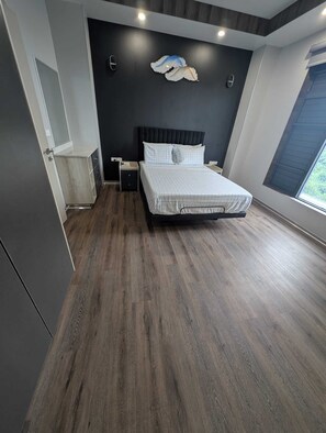 A modern bedroom with a large bed, dark wood flooring, a dark wall with a decorative winged design, and a window with a view.
