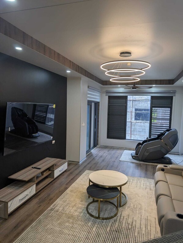 A modern living room with a flat-screen TV, reclining chairs, a round coffee table, and a ceiling light fixture.