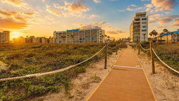 A wooden boardwalk leading to a beachfront hotel with multiple floors and balconies, surrounded by vegetation and palm trees.