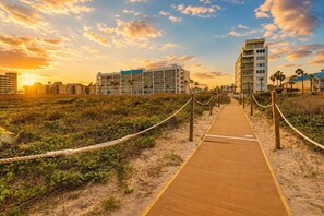 Un paseo marítimo de madera que conduce a un hotel frente a la playa con varios pisos y balcones, rodeado de vegetación y palmeras.