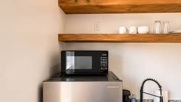 Un refrigerador de acero inoxidable con un microondas negro encima, un estante de madera arriba con una planta en maceta, y utensilios de cocina como una tetera y un rollo de toallas de papel.