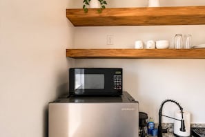 Un refrigerador de acero inoxidable con un microondas negro encima, un estante de madera arriba con una planta en maceta, y utensilios de cocina como una tetera y un rollo de toallas de papel.