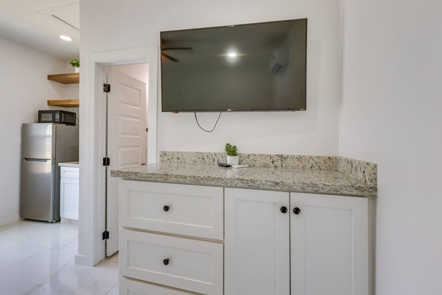 A kitchen with white cabinets, a granite countertop, a stainless steel refrigerator, a microwave, and a flat-screen TV mounted above the counter.