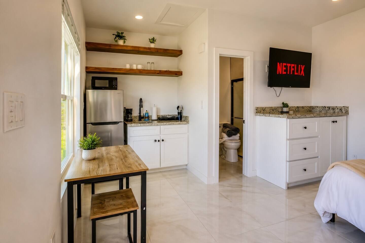 A modern kitchen with a refrigerator, microwave, and wooden shelves. There is a dining table with chairs and a TV mounted on the wall displaying 'NETFLIX'.
