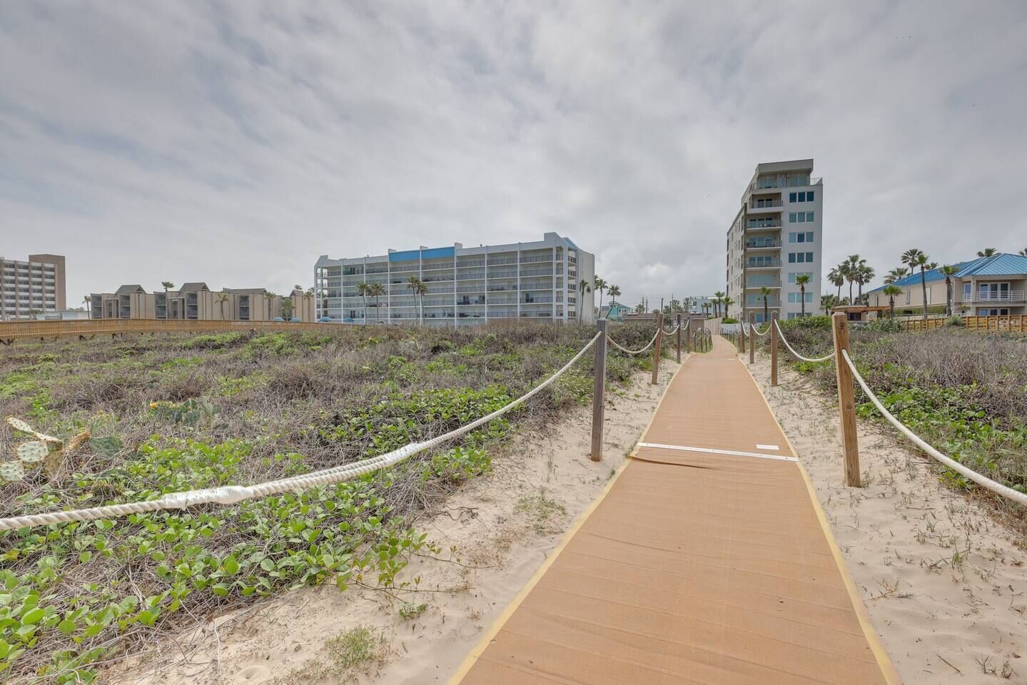 Un sendero arenoso que lleva a un hotel frente al mar con varios pisos y balcones, rodeado de vegetación y otros edificios.