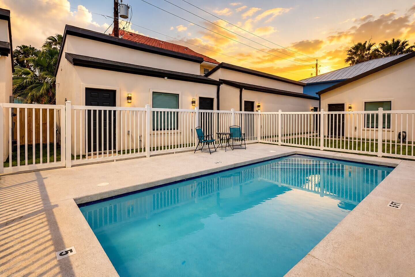 A swimming pool with a white fence, a covered patio area, and a building with a red roof.