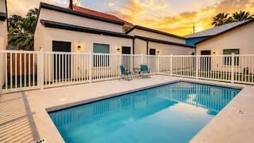 A swimming pool with a white fence, a covered patio area, and a building with a red roof.