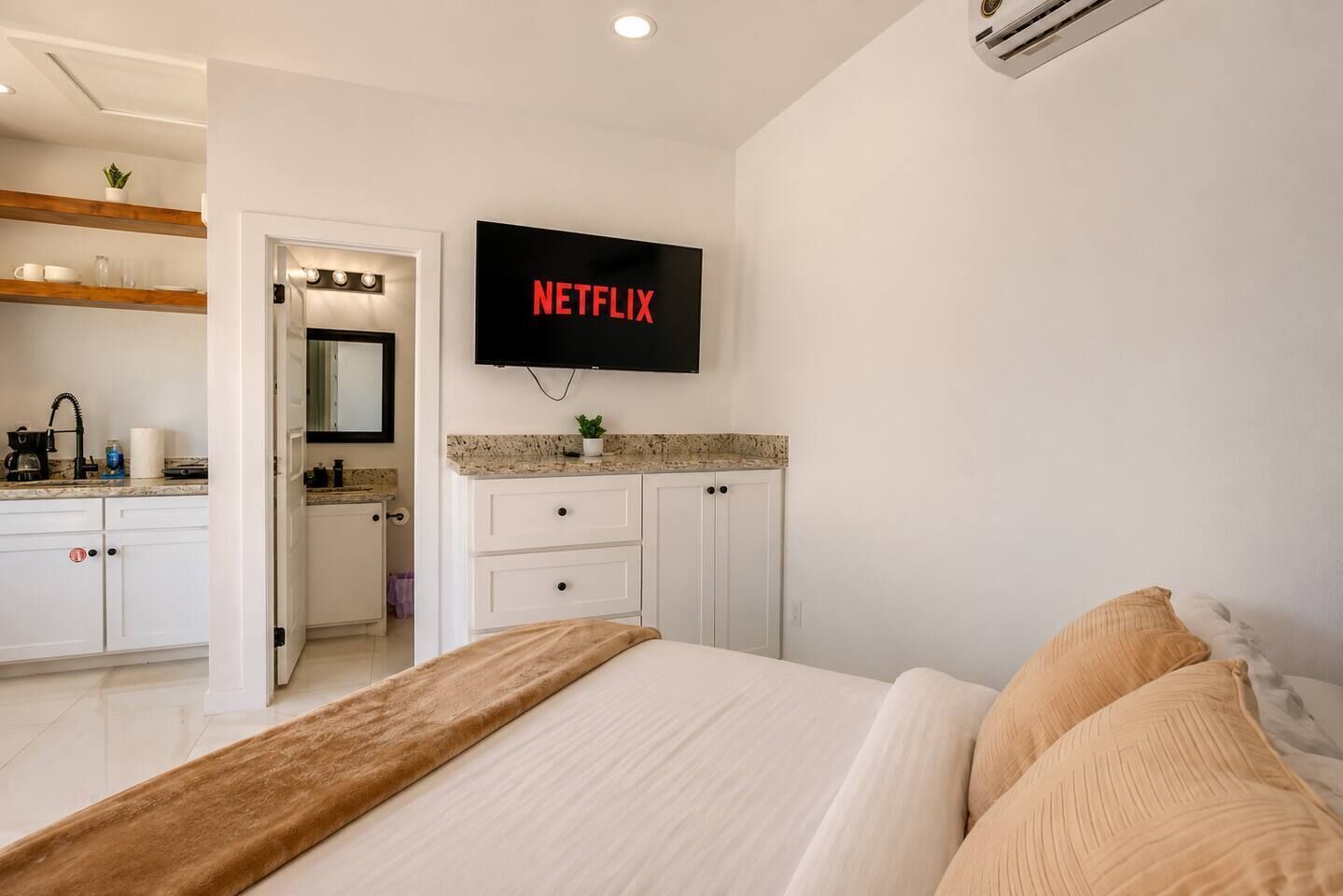 A hotel room with a bed, a television mounted on the wall, a bathroom with a mirror, and a kitchenette area.