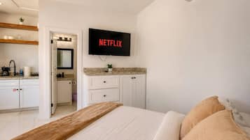 A hotel room with a bed, a television mounted on the wall, a bathroom with a mirror, and a kitchenette area.