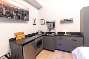 A compact kitchen with a built-in oven, a sink, and a small countertop. There is a wooden cutting board on the counter, and a cityscape painting hangs on the wall. - Spruce Villa - 2nd Floor Queen Studio (Philadelphia)