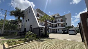 Edifício residencial de vários andares com entrada protegida por portão, calçada pavimentada e área de jardim ajardinada.