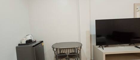 Pequena sala de estar com TV de tela plana em suporte, mesa pequena com duas cadeiras, geladeira compacta e lixeira.