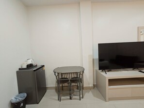 A small living area with a flat-screen TV on a stand, a small table with two chairs, a compact refrigerator, and a trash bin.