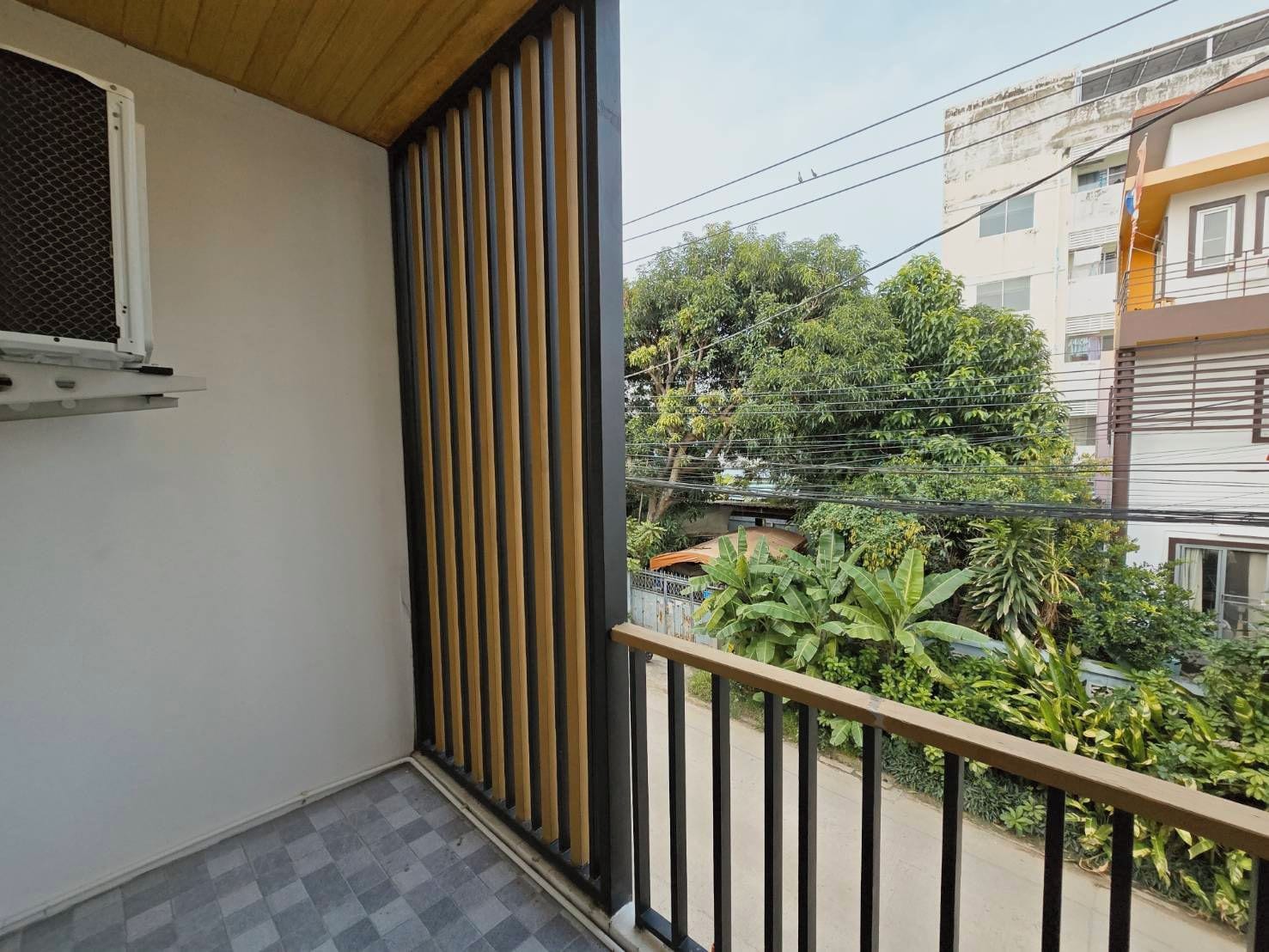 A balcony with a checkered floor, a window with a mesh screen, and a view of neighboring buildings and greenery.