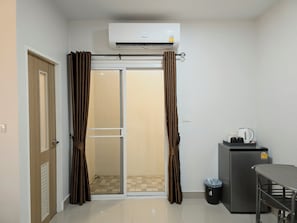 A room with a sliding glass door, an air conditioning unit, a small refrigerator, and a table with a kettle. - Hada House near Yanhee Hospital (Bangkok)