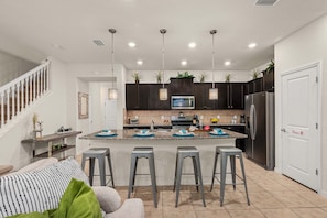 A modern kitchen with a central island, dark cabinets, stainless steel appliances, and a staircase leading to another level. - 5BR Solterra Escape w/ Private Pool & Themes (Davenport)