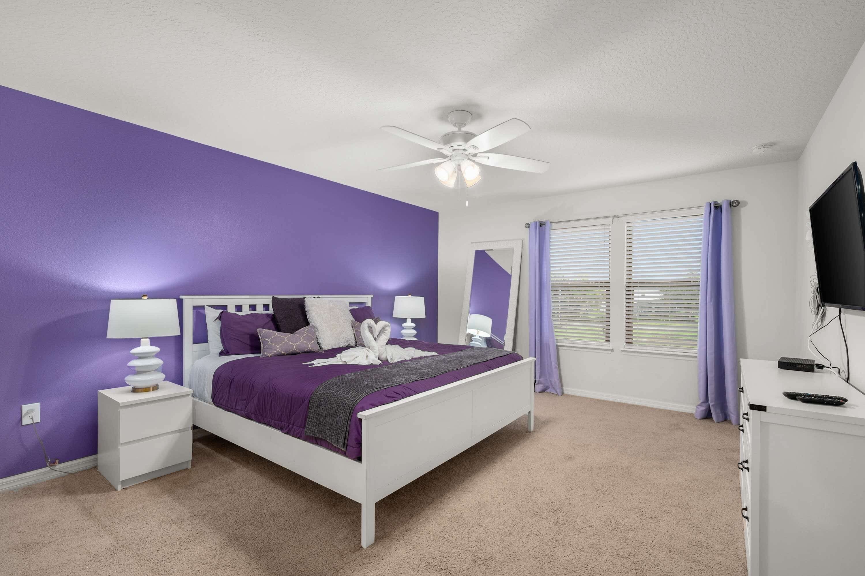 A bedroom with a white bed, purple walls, a ceiling fan, and a window with blinds.