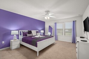 Ein Schlafzimmer mit einem weißen Bett, violetten Wänden, einem Deckenventilator und einem Fenster mit Jalousien.