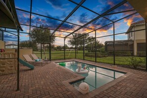 A swimming pool with a slide and inflatable toys, surrounded by a brick patio and a glass enclosure with a view of a sunset. - 5BR Solterra Escape w/ Private Pool & Themes (Davenport)