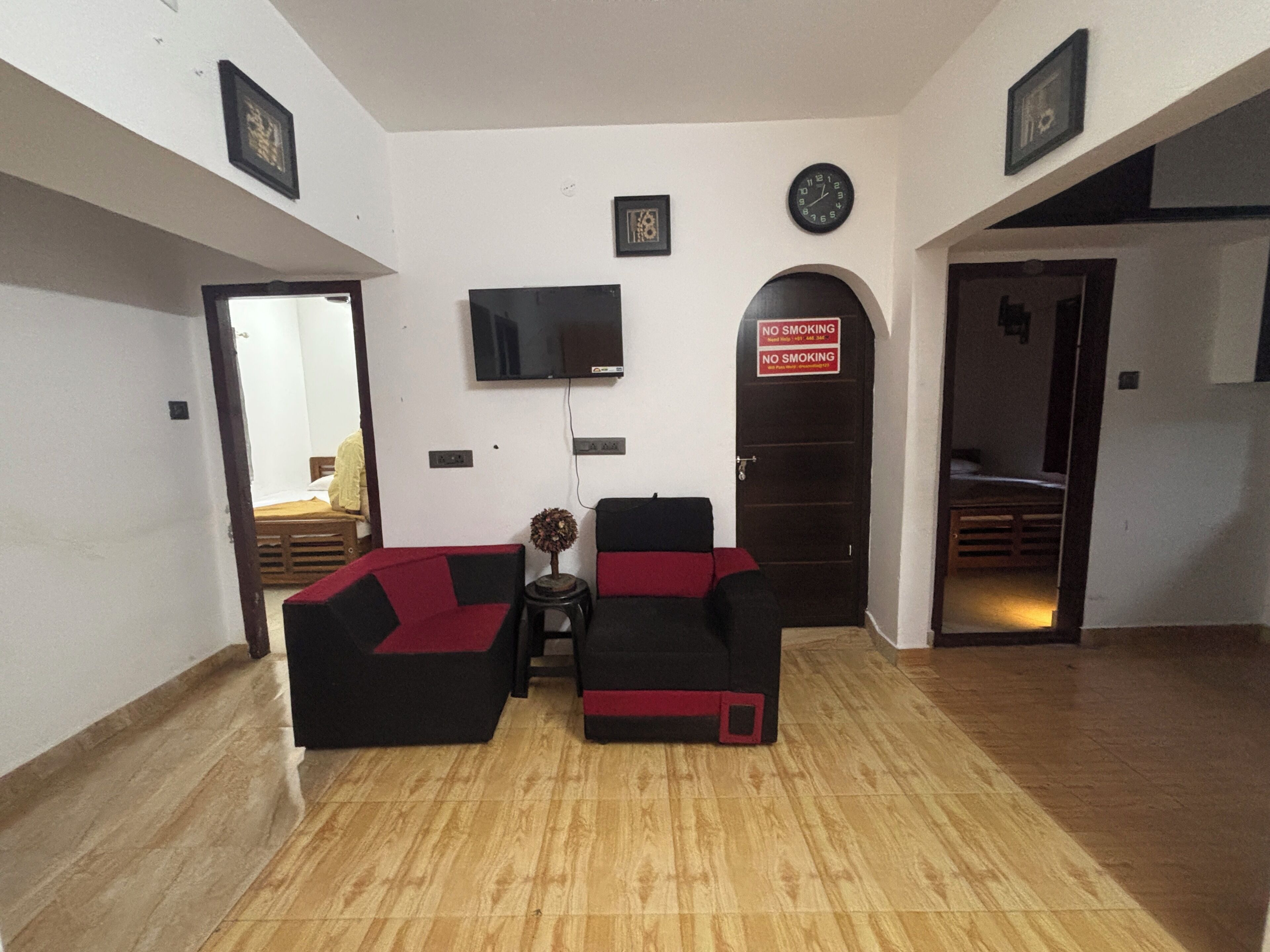 A living room with a flat-screen TV mounted on the wall, a sofa set, a small table with a vase, and a door with a 'No Smoking' sign.