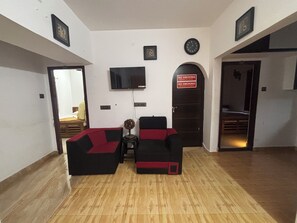 A living room with a flat-screen TV mounted on the wall, a sofa set, a small table with a vase, and a door with a 'No Smoking' sign.