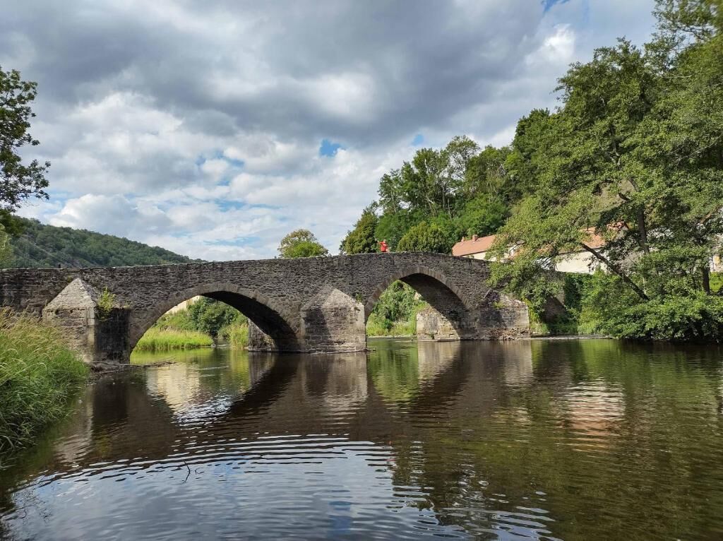 A stone bridge over a calm river with trees and a building on the right.