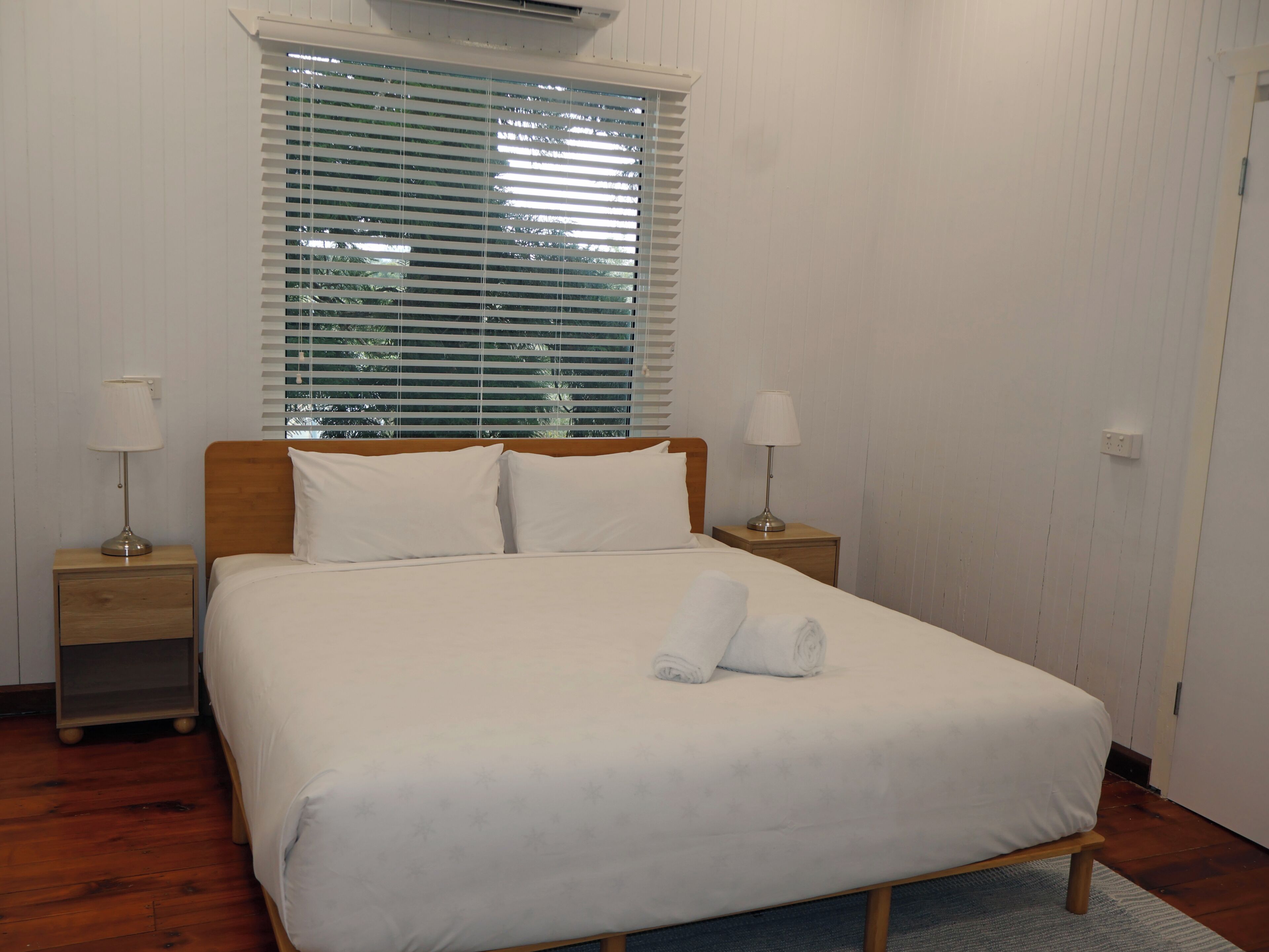 Una cama bien tendida con sábanas blancas y una toalla enrollada, cabecera de madera, dos lámparas de noche y una ventana con persianas.