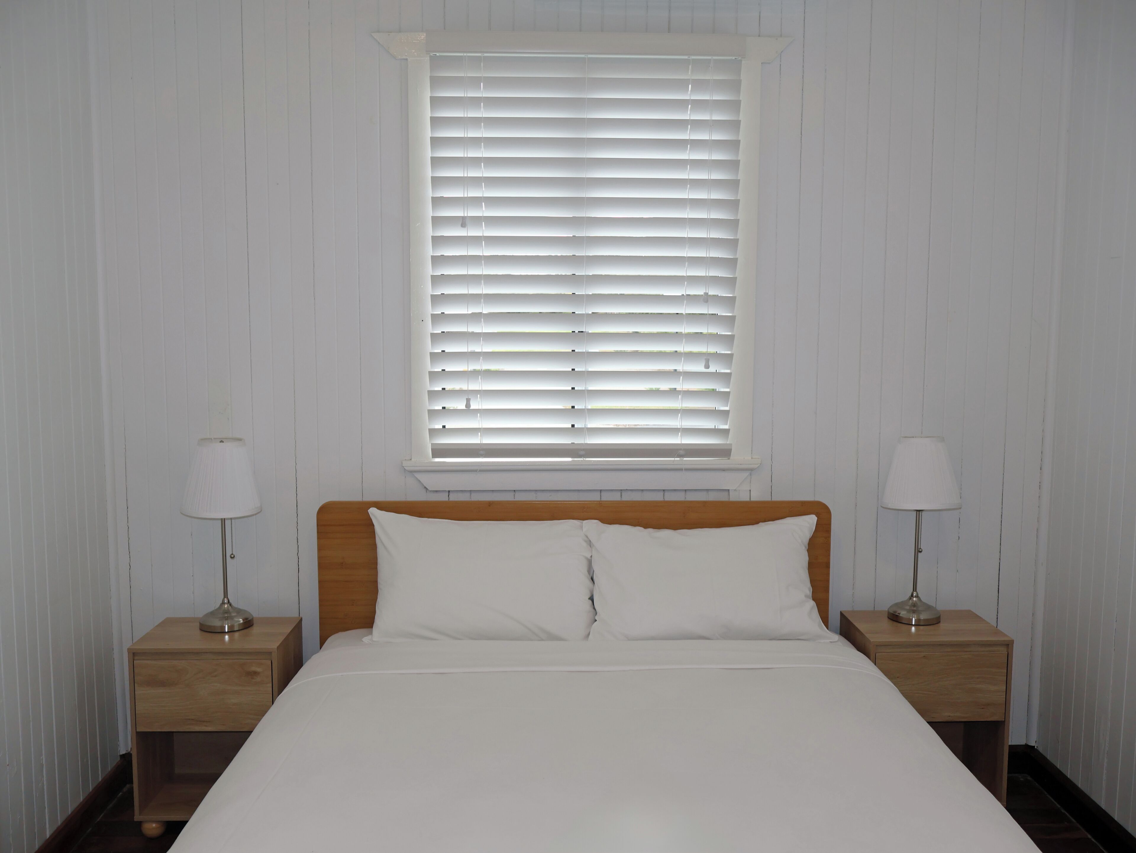 A bedroom with a wooden headboard, white bedding, two bedside tables with lamps, and a window with blinds.