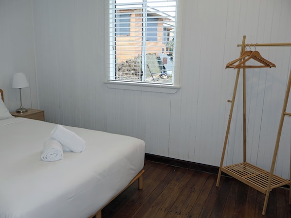 A hotel room with a bed, a wooden hanger rack, a window with blinds, and a view of buildings outside. - Gympie Cottage (Gympie)