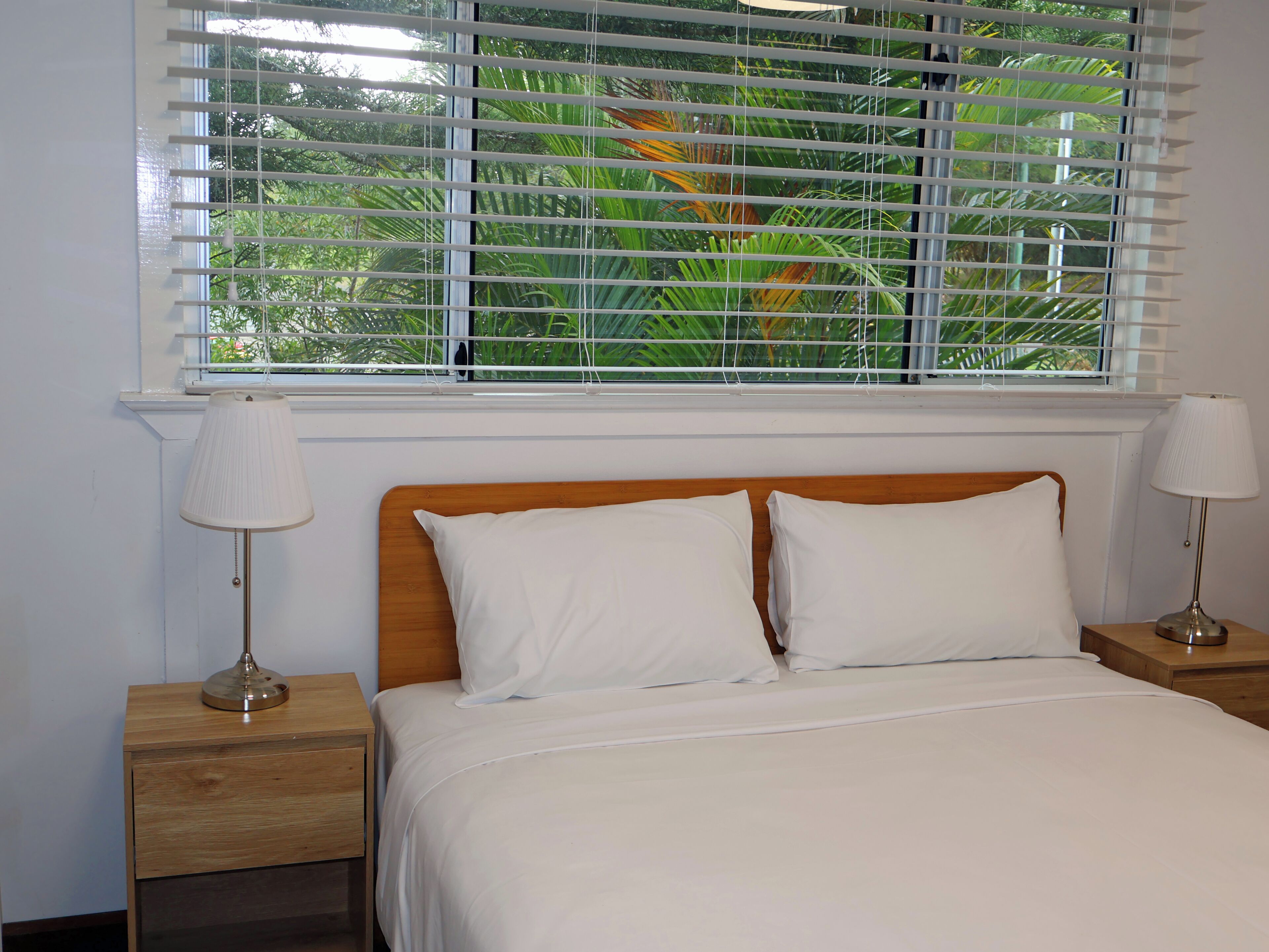 Een bed met wit beddengoed, twee bedlampjes en een raam met zonwering.