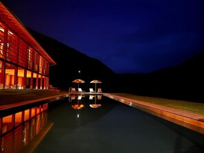Una piscina con sillones y sombrillas, rodeada por un edificio con grandes ventanales y un cielo nocturno.