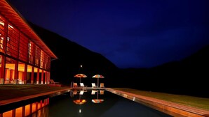 Ein Schwimmbecken mit Liegestühlen und Sonnenschirmen, umgeben von einem Gebäude mit großen Fenstern und Nachthimmel.
