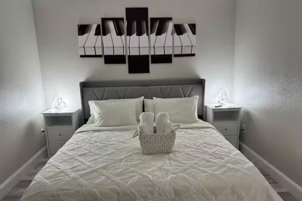 Un lit bien fait, avec une couette blanche et des oreillers, une table de chevet avec une lampe, et une œuvre d’art abstraite accrochée au mur.