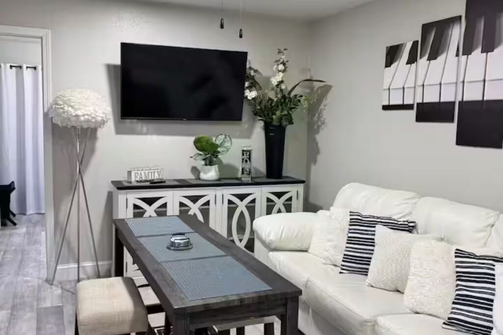 Uma sala de estar moderna com um sofá branco, uma mesa de centro, uma TV fixada na parede e objetos decorativos, incluindo um vaso com flores.