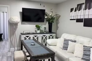 Un salón moderno con un sofá blanco, una mesa de centro, un televisor montado en la pared y objetos decorativos, incluyendo un jarrón con flores.