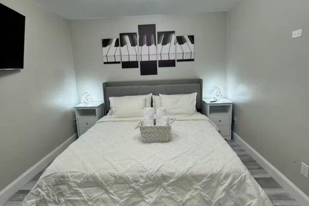 Cama bem arrumada com edredom branco e almofadas, mesa de cabeceira com abajur e uma obra de arte abstrata na parede.