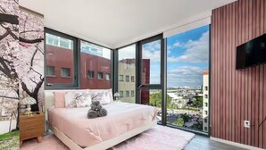 Quarto com uma cama grande, mesa de cabeceira, TV fixada na parede e vista da cidade por grandes janelas.