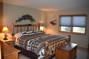 Un dormitorio con cabecera de madera, cama con colcha estampada, dos lámparas de noche, una ventana con persianas y un estante en la pared.