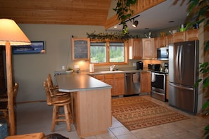 A modern kitchen with wooden cabinets, stainless steel appliances, a large island with bar stools, and a view of the outdoors through the windows. - Mountainside Georgetown Lake Cabin with Sweeping Lake & Pintler Mountain Views (Anaconda)