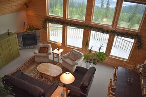 A cozy cabin interior with a fireplace, comfortable seating, and large windows overlooking a forest. - Mountainside Georgetown Lake Cabin with Sweeping Lake & Pintler Mountain Views (Anaconda)
